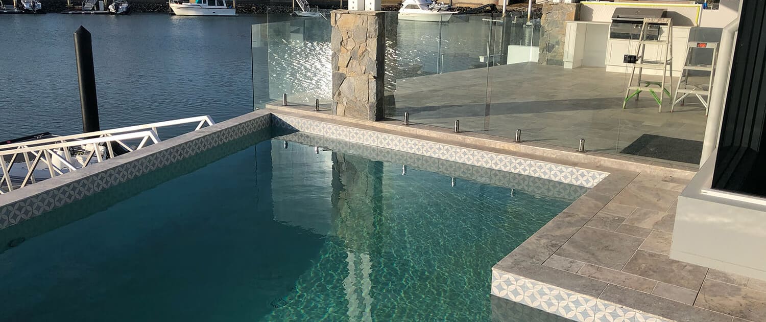 A luxurious outdoor swimming pool, crafted by renowned Pool Builders Brisbane, features intricate tiled borders and clear blue water. Surrounding the pool is a spacious patio with glass railing. Beyond the pool, a serene waterfront is visible with houses and boats docked along the shoreline.