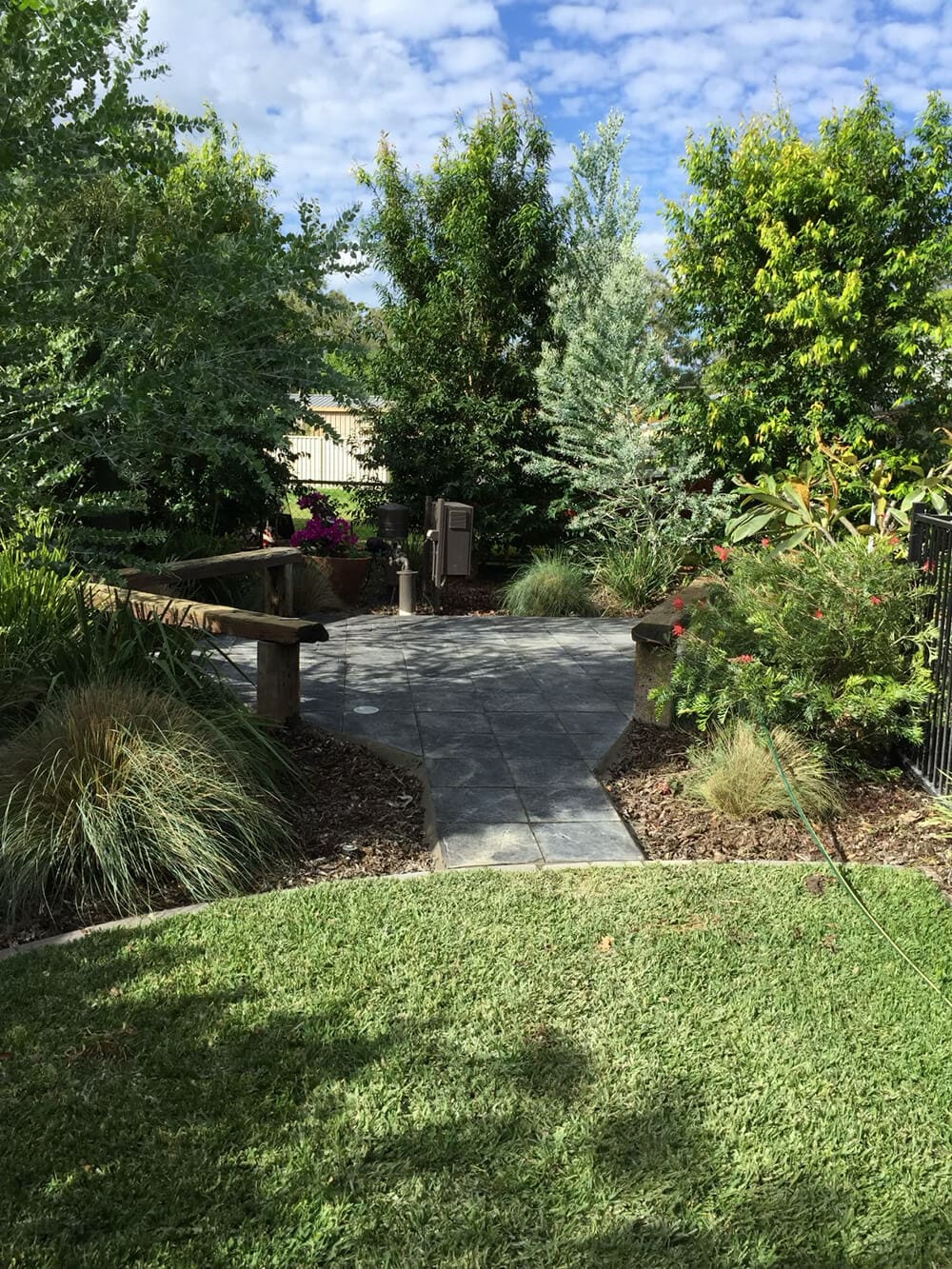 A serene backyard garden, designed by Adella Pools, features a stone-paved path leading to a wooden bench surrounded by lush greenery. Various trees and shrubs, along with neatly trimmed grass and vibrant plants, create a tranquil and inviting atmosphere under a partly cloudy sky.