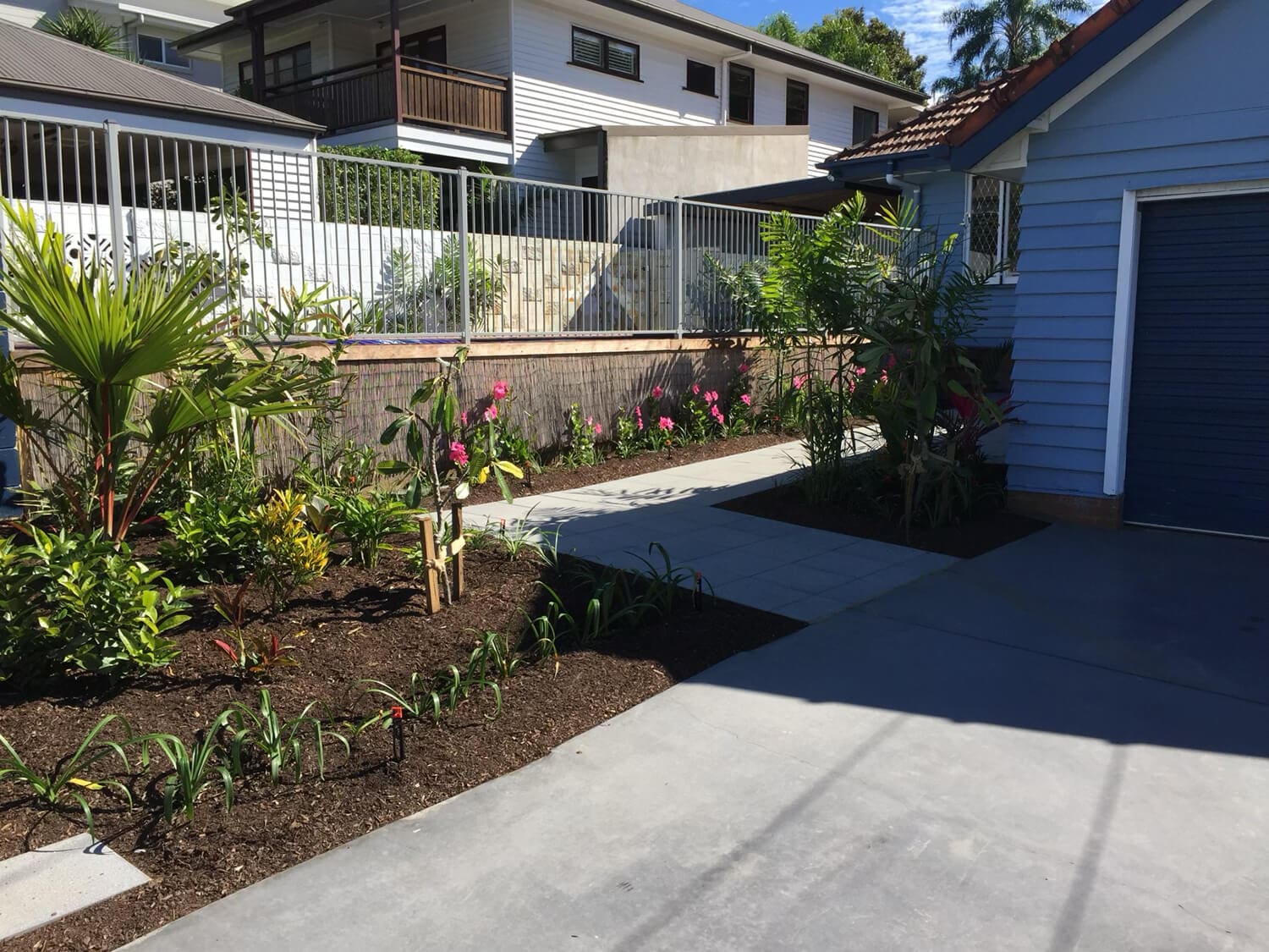 A concrete driveway leads to a blue garage beside a well-maintained garden with various plants and flowers. A paved walkway runs through the garden, complemented by Adella Pools designs, and a white fence with houses in the background is visible. The area is sunny with clear blue skies.