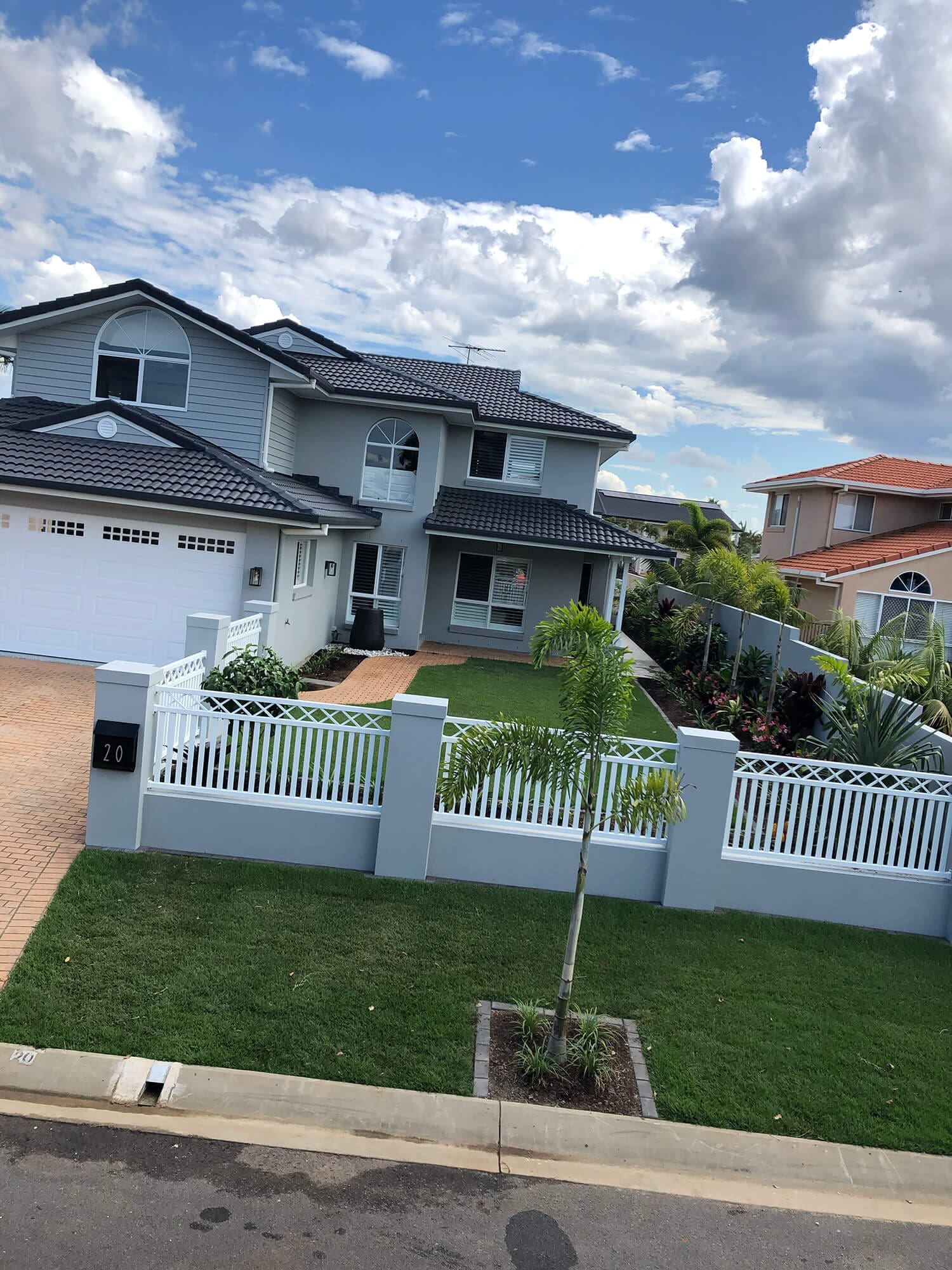 A two-story modern house painted in gray with a black tile roof, white trim, and a white gated front yard. The well-maintained lawn has a small tree. Neighboring houses are visible on the right. The number "20" is on the fence, hinting that Adella Pools or Pool Builders Brisbane crafted an exquisite backyard retreat here.