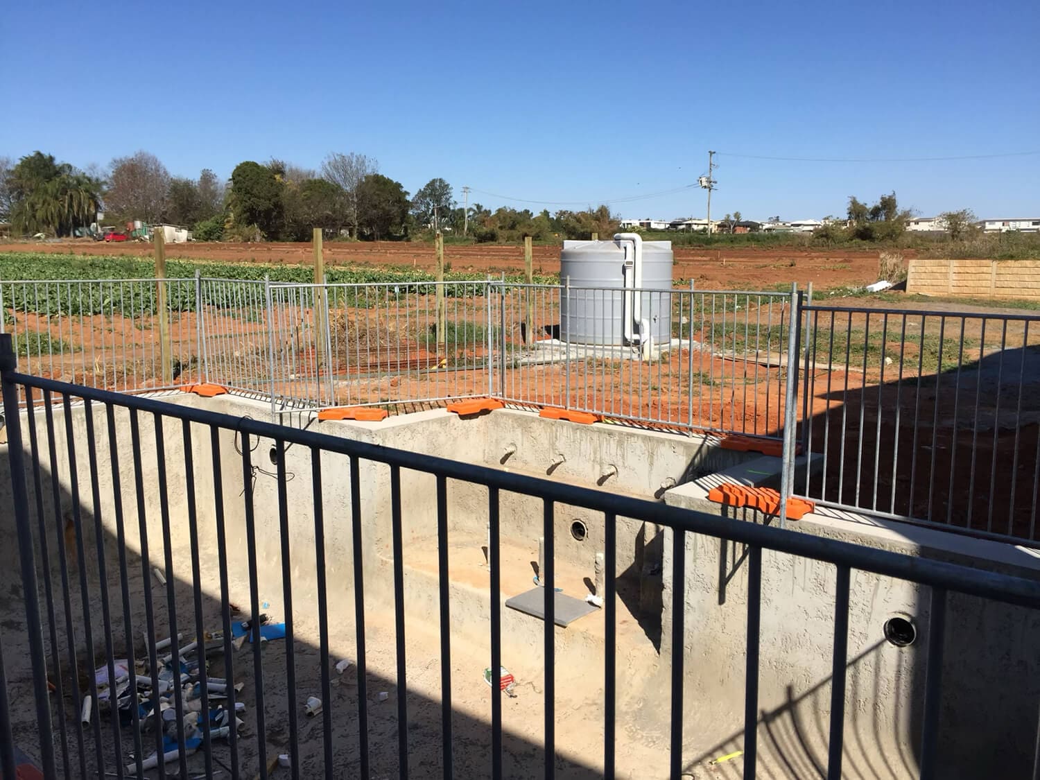 A construction site for an in-ground swimming pool, managed by Pool Builders Brisbane, is surrounded by a temporary metal fence. The pool bed is unfinished, with tools and construction materials scattered around. In the background, there's an open field, a small building, and a clear blue sky.