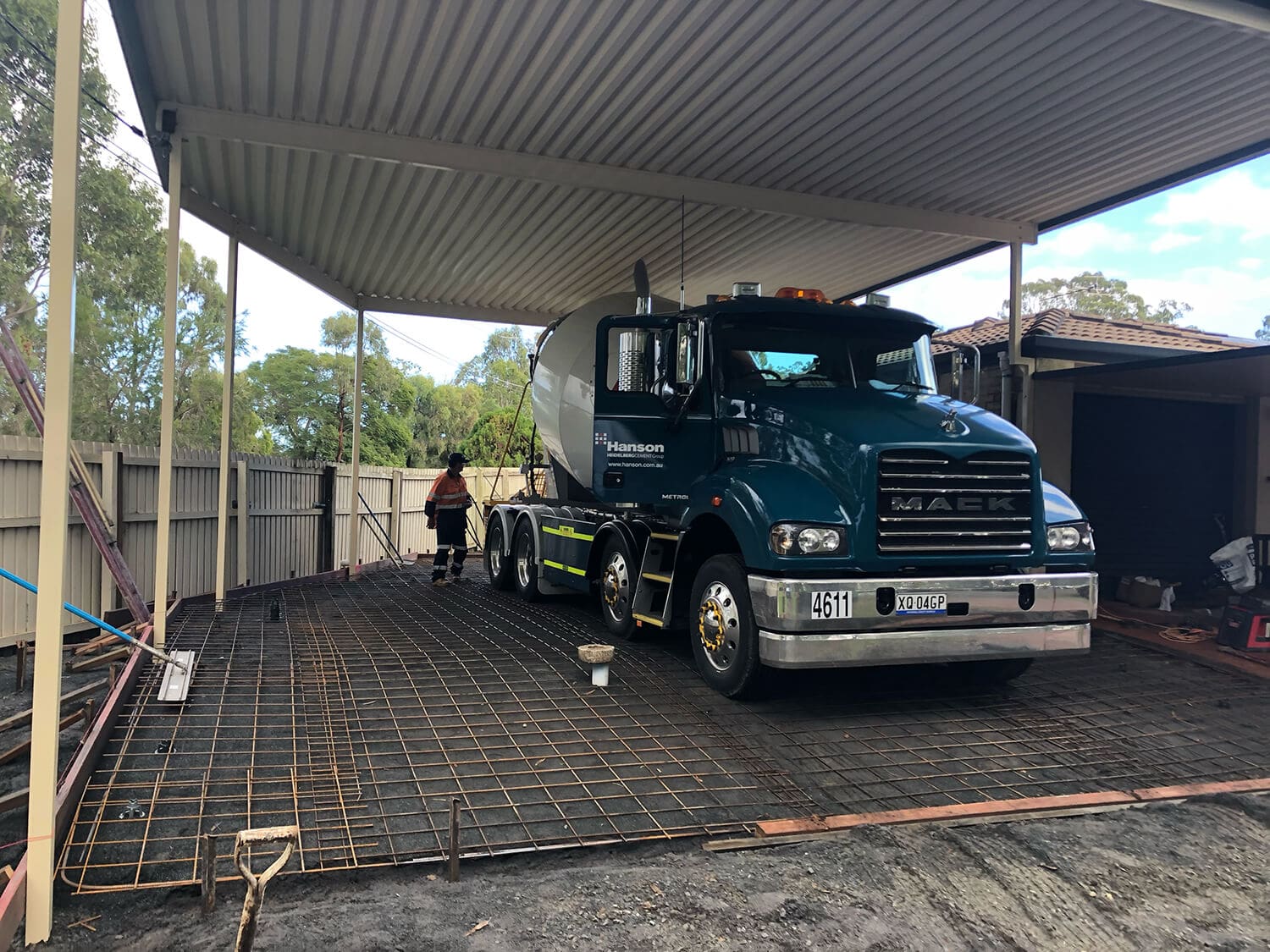 A truck with the label "Hanson" is parked under a carport on a rebar grid, likely preparing for concrete pouring. Two workers wearing safety gear are seen at the rear of the truck. Trees and buildings are visible in the background, indicative of Pool Builders Brisbane hard at work.