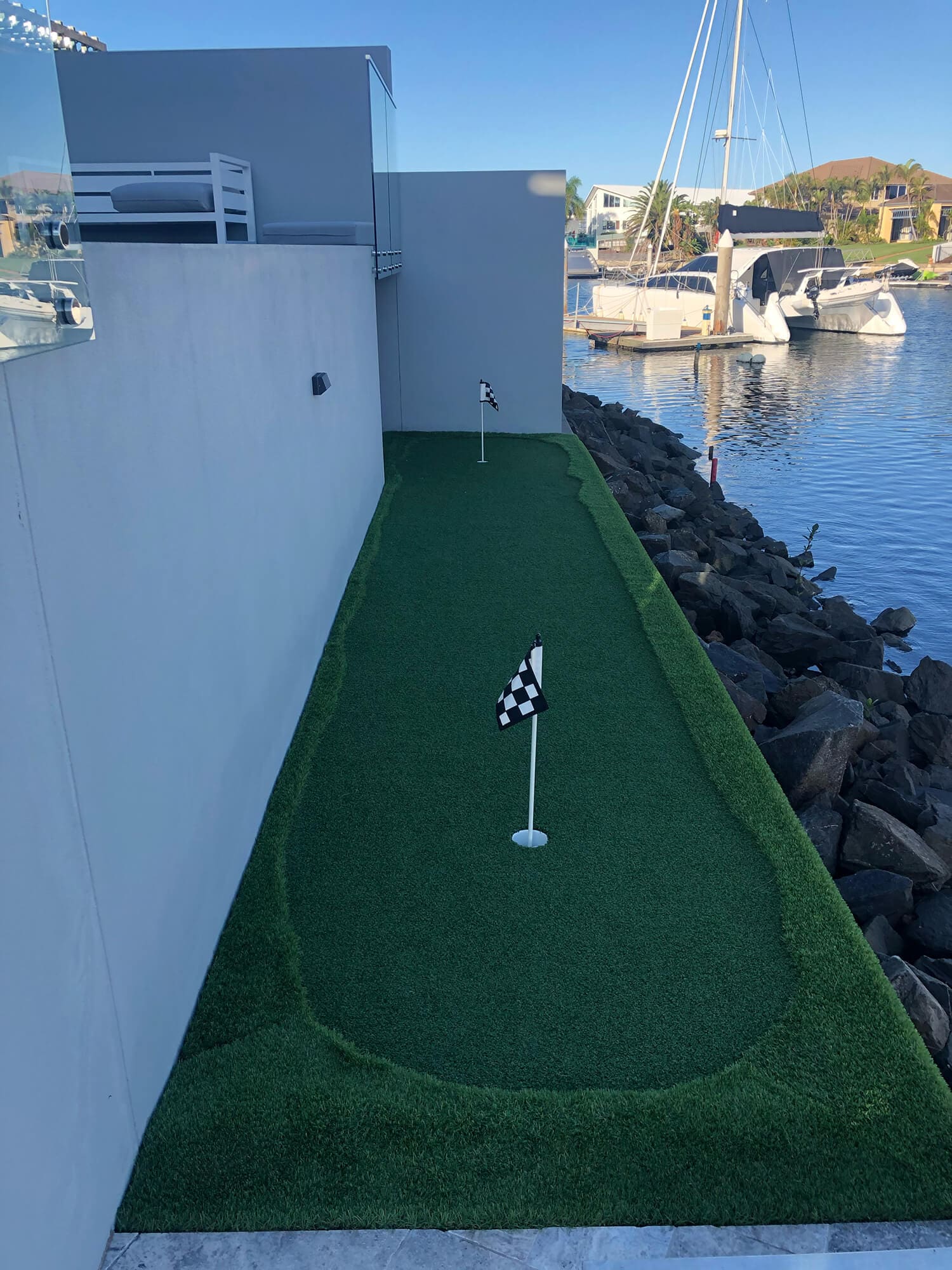 A rooftop mini-golf course with two checkered flag holes is situated beside a modern gray building designed by Pool Builders Brisbane. The course overlooks a calm marina with a docked sailboat and adjacent homes in the background, all bathed in bright, sunny weather.