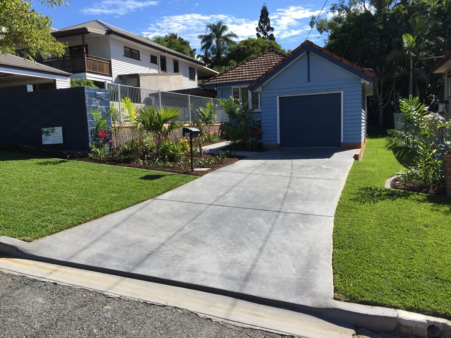 A small blue house with a brown-tiled roof is seen from the street. A concrete driveway leads up to a single-car garage. The house, perfect for those consulting Pool Builders Brisbane, is surrounded by a tidy lawn and various plants. Neighboring houses are visible in the background under a clear blue sky.