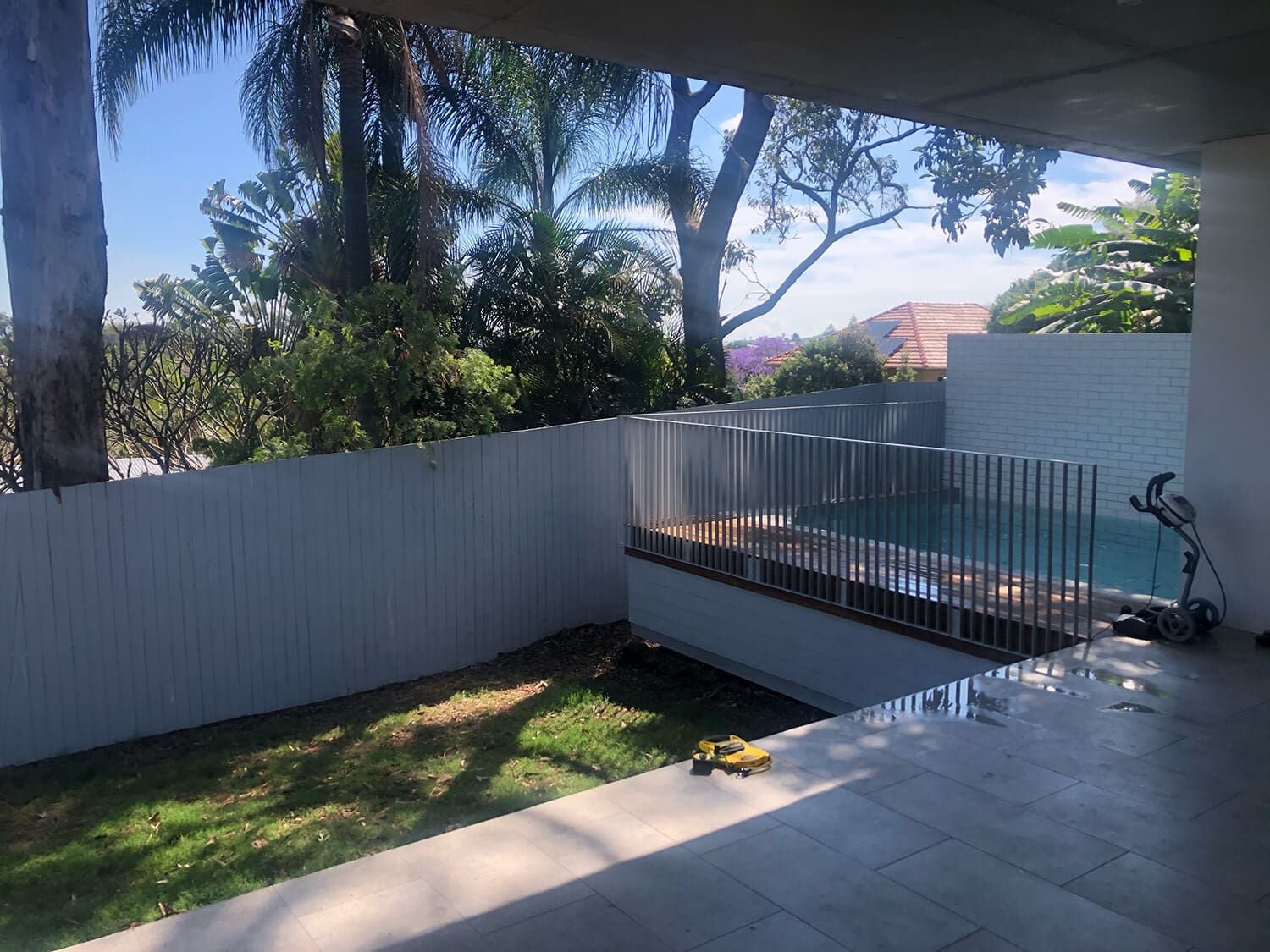 A backyard features a small grassy area, a tiled patio, and a raised swimming pool with a metal fence crafted by Pool Builders Brisbane. A green scooter and a yellow toy lie on the ground. The scene is framed by trees and shrubbery with a clear sky overhead.