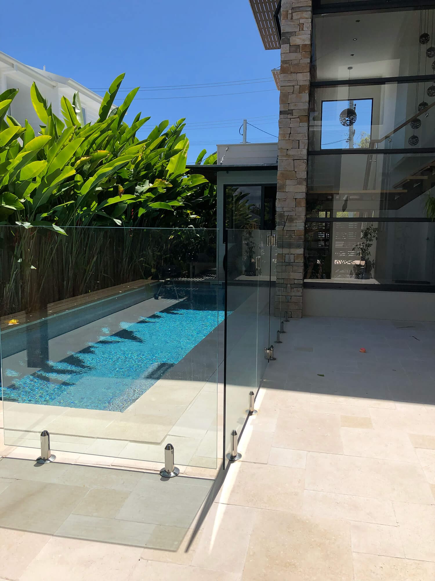 A modern outdoor pool area features a rectangular swimming pool surrounded by a glass fence. The stone patio beside the pool leads to a contemporary house with large glass windows. Lush green tropical plants are visible in the background, showcasing the expertise of Pool Builders Brisbane.