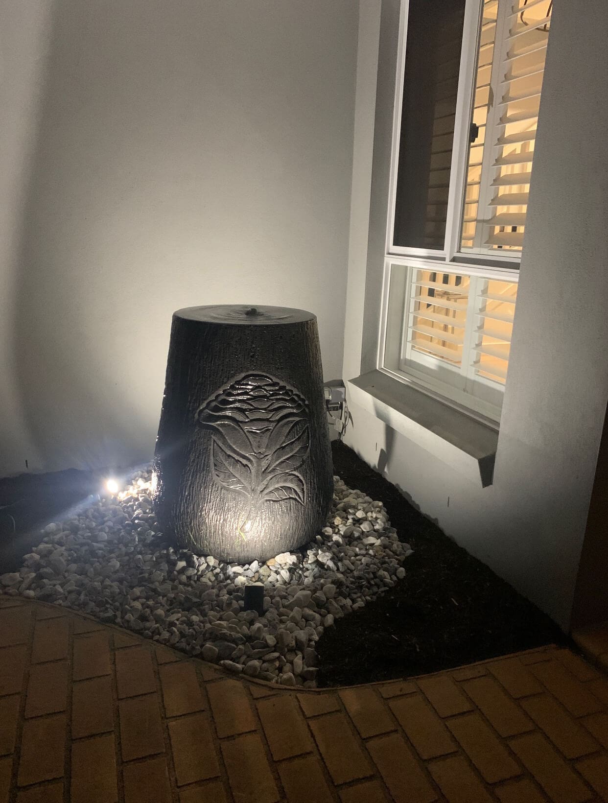 A lit black tree stump planter with an engraved leaf design, surrounded by small white stones and illuminated by a spotlight, is placed next to a white house wall and a window with wooden shutters. Brick pavement, often seen in projects by Pool Builders Brisbane, is visible in the foreground.