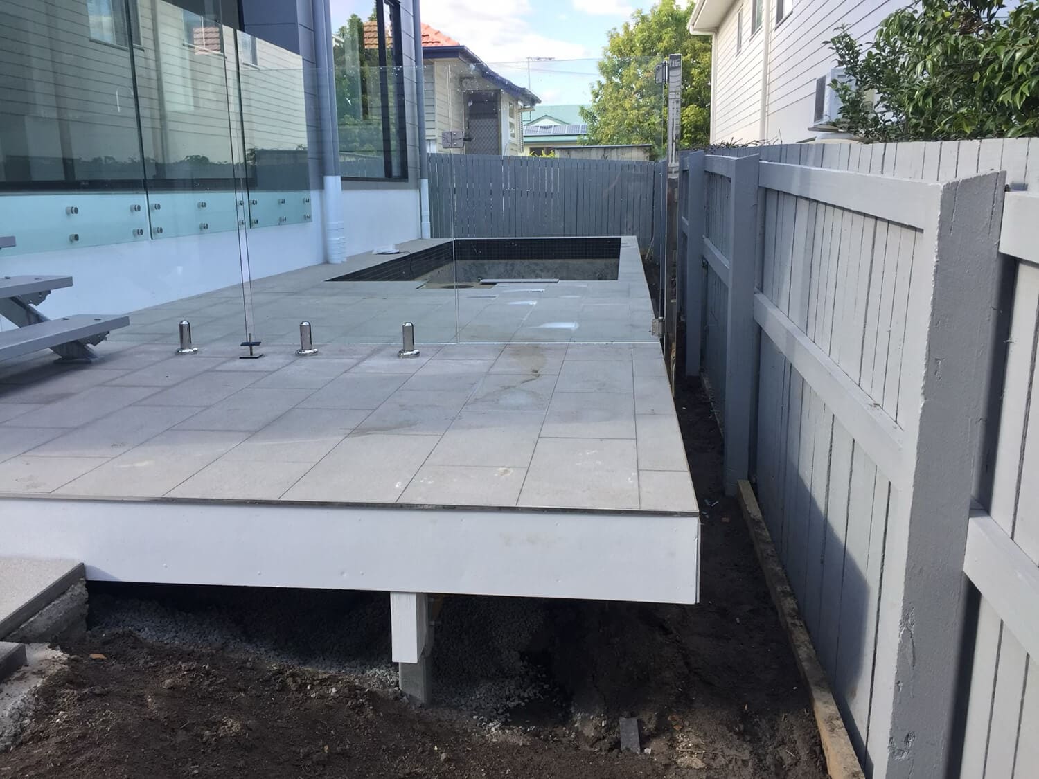 A modern backyard under construction features a raised tiled patio beside a glass railing, with an empty pool area being expertly crafted by Pool Builders Brisbane. Surrounding the patio is a partially built fence, and construction materials are scattered on the ground below.