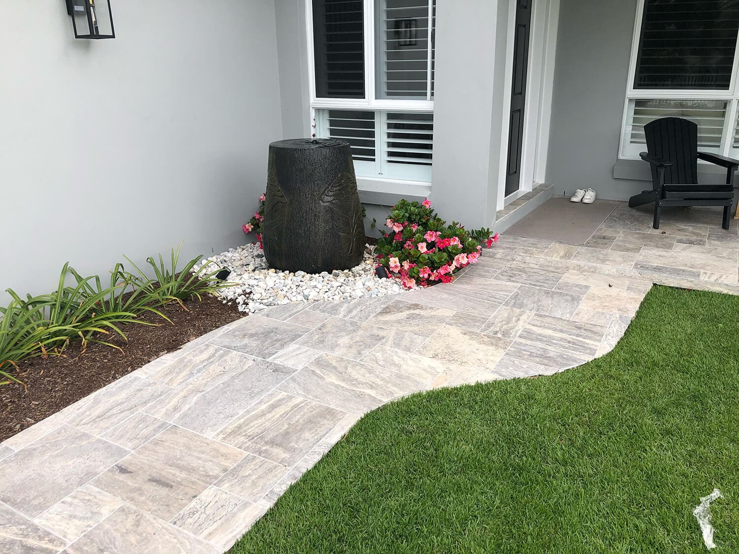 A neatly landscaped patio area with stone tiles, bordered by a green lawn. The corner features a black stone water fountain, surrounded by white pebbles and pink flowers. There is a black chair by the entrance, and a pair of white shoes near the door. For enhancements like this, consult Pool Builders Brisbane.