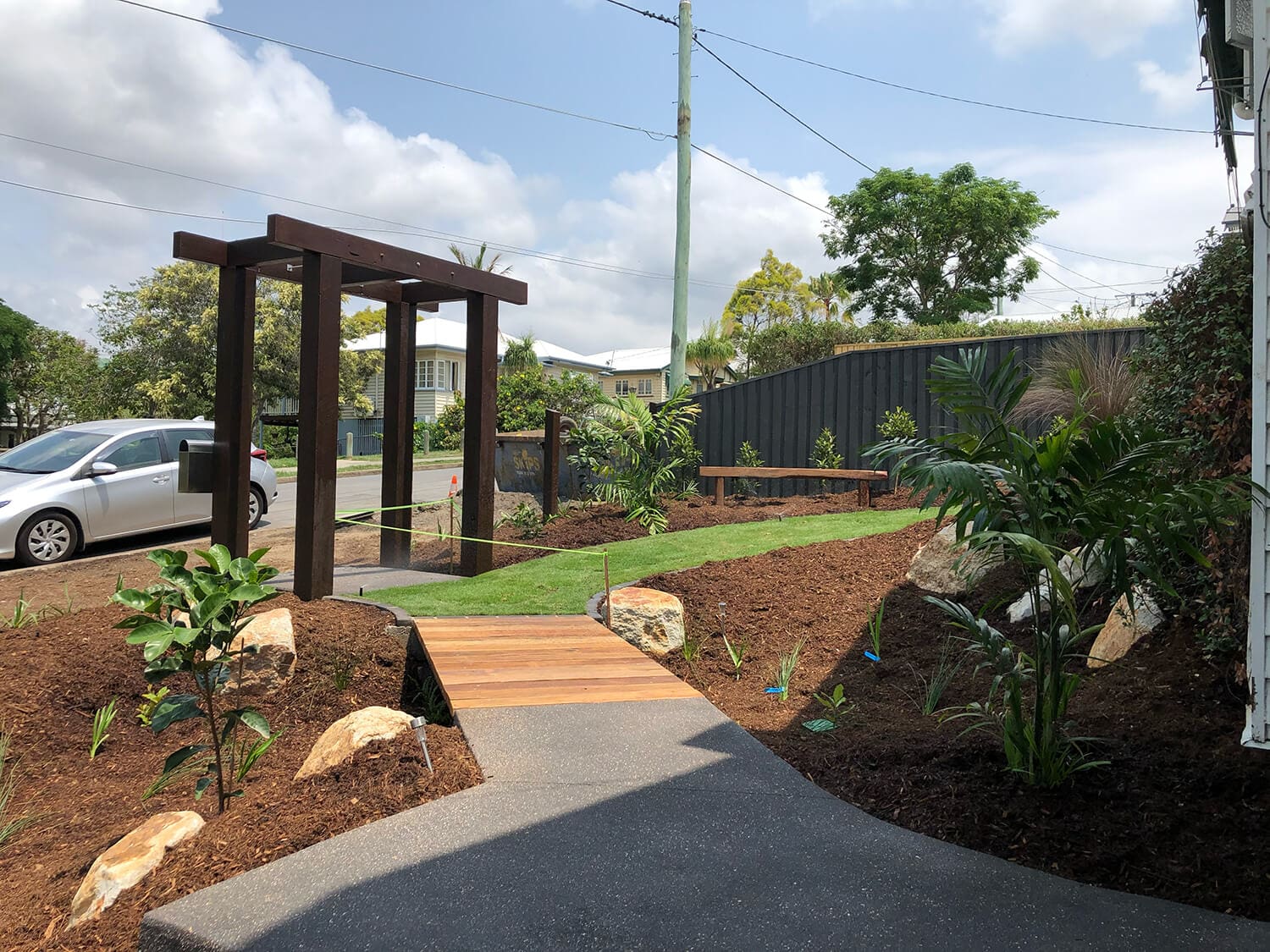 A newly landscaped garden with a pathway leading to a wooden arbor. The area features freshly planted shrubs, young trees, and mulch beds. A car is parked on the left side near the entrance. A black fence runs along the back, with houses and a cloudy sky in the distance—a perfect scene for Pool Builders Brisbane to craft an inviting oasis.