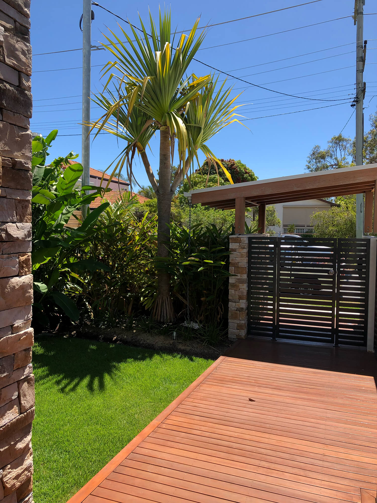 A lush backyard with a wooden deck, a neatly manicured lawn, and a tall palm tree beside a stone column. In the background, Pool Builders Brisbane showcase their artistry with green vegetation and a modern metal gate flanked by stone pillars under a clear blue sky. Power lines are visible overhead.