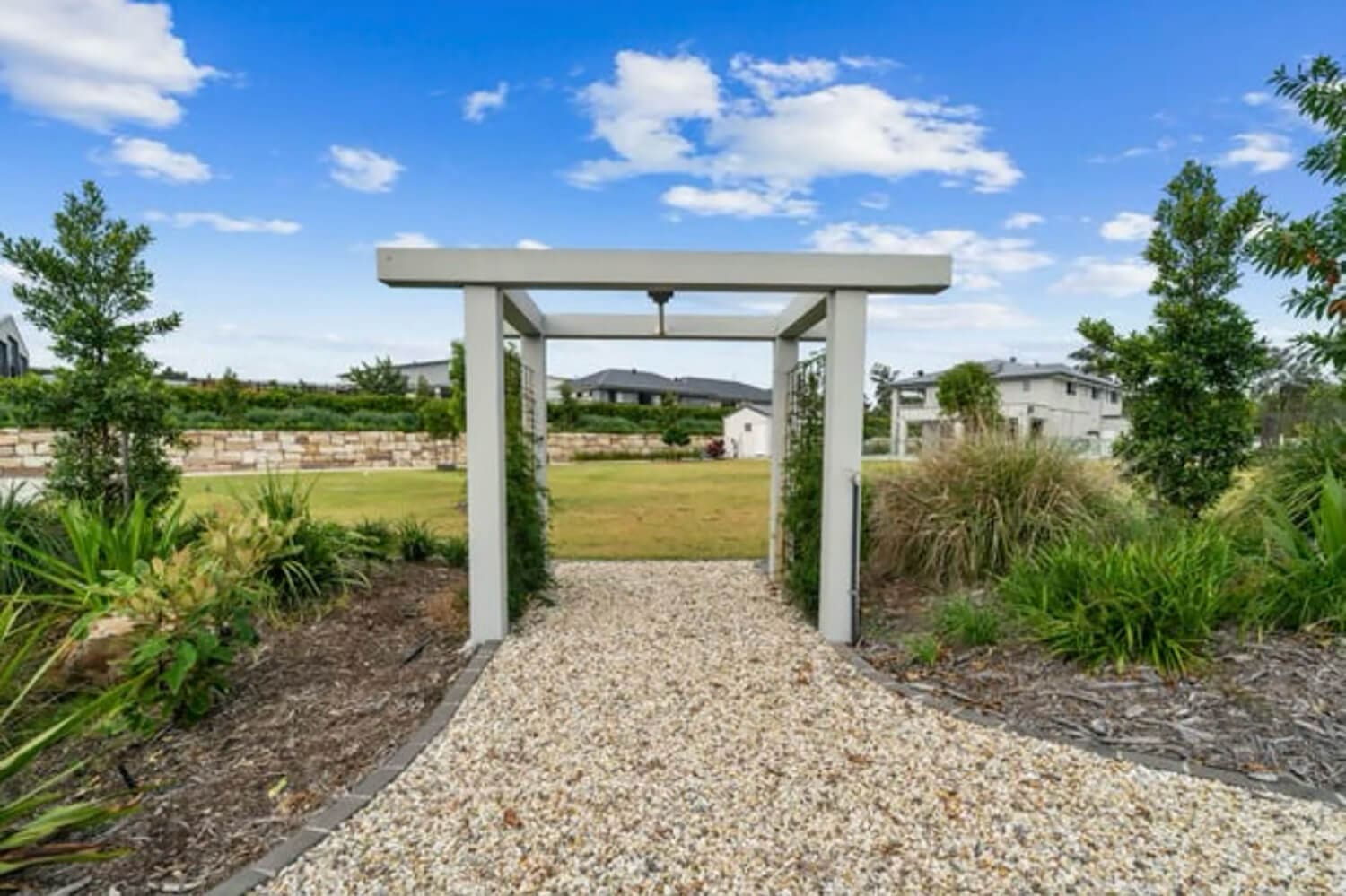 A gravel pathway leads through a white wooden garden archway surrounded by green shrubs and trees, opening up to a spacious grassy area with buildings, where Pool Builders Brisbane have crafted an inviting oasis under the bright blue sky.