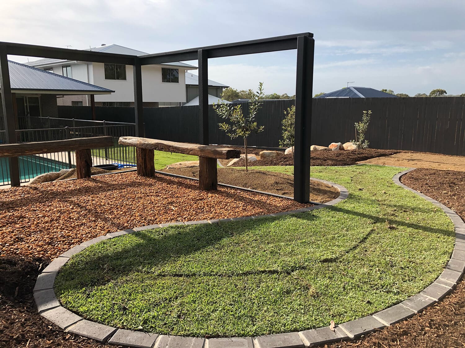 A backyard with a newly landscaped area featuring a rectangular black frame structure, a section with green grass bordered with pavers, a wood chip area with wooden beam accents, young trees, and a nearby pool expertly crafted by Pool Builders Brisbane and fenced with a black metal fence.