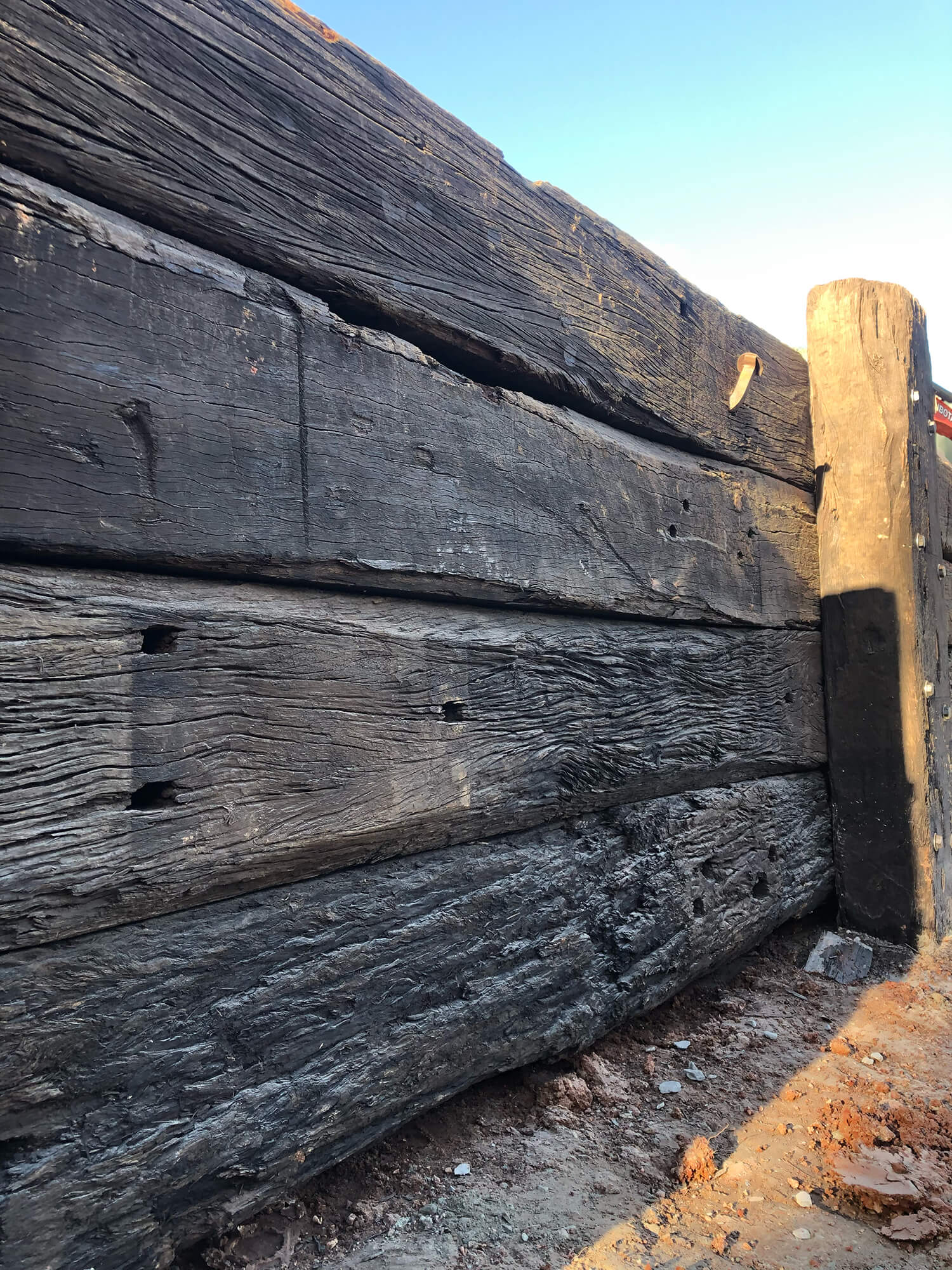 A close-up of a weathered wooden retaining wall with large, rough-hewn timber beams stacked horizontally. The wood appears aged, with visible cracks, holes, and texture. Crafted to perfection by expert Pool Builders Brisbane, the wall is set against a clear blue sky, with dirt and debris at its base.