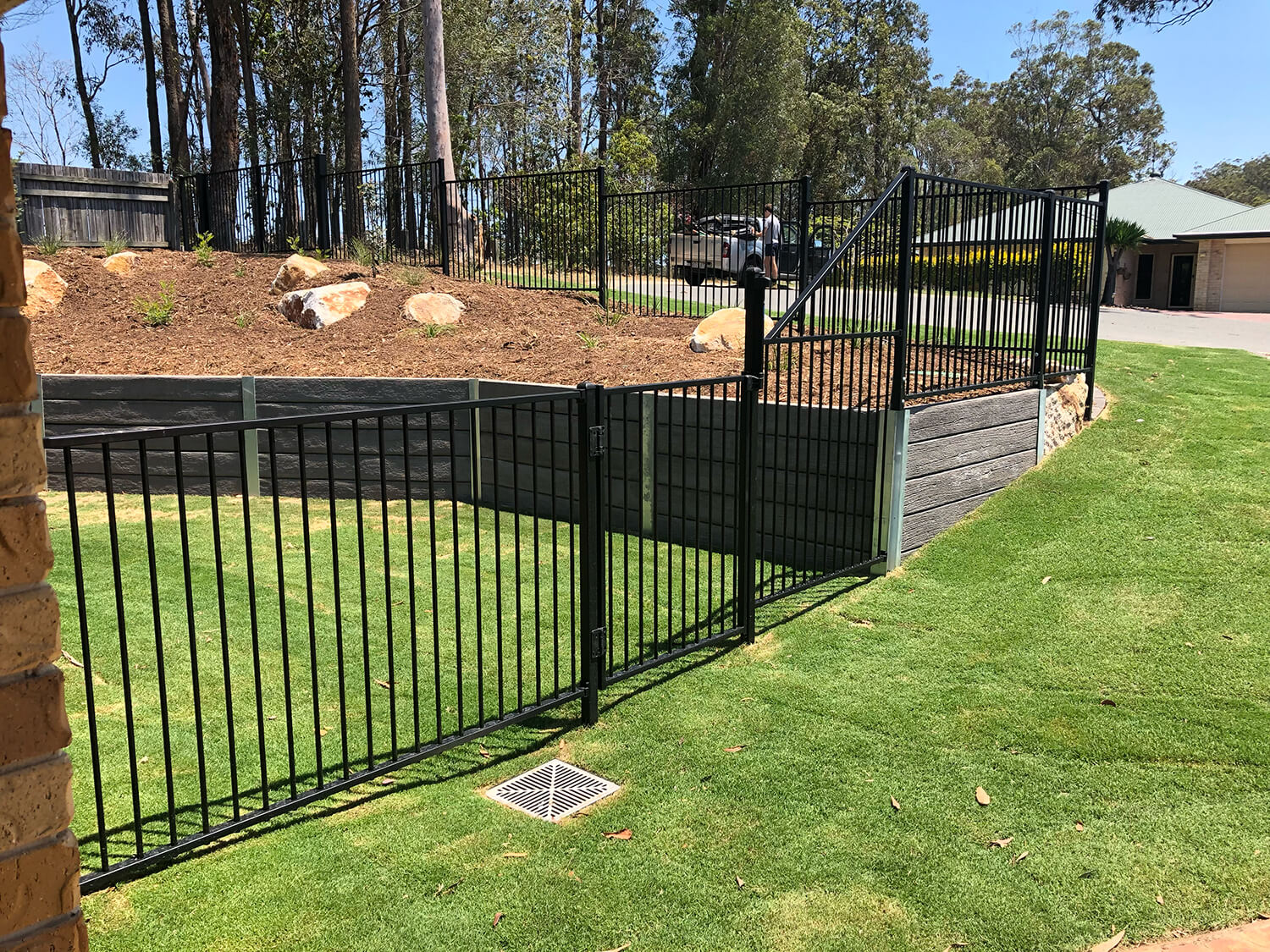 A black metal fence encloses a grassy yard area with a gate. Beyond the fence, there is an elevated section of land with rocky landscaping under tall trees, perfect for creative touches by Pool Builders Brisbane. The background features more greenery and a residential building with a green roof.