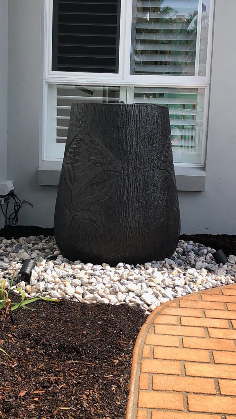 A sculpted black stone vase is placed in a garden bed filled with small white pebbles. The vase has floral engravings and is positioned in front of a window with white shutters. The area is bordered by a brown brick pathway.